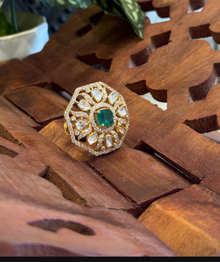 An ornate gold ring with a central green emerald and surrounding clear stones, displayed on a carved wooden surface.